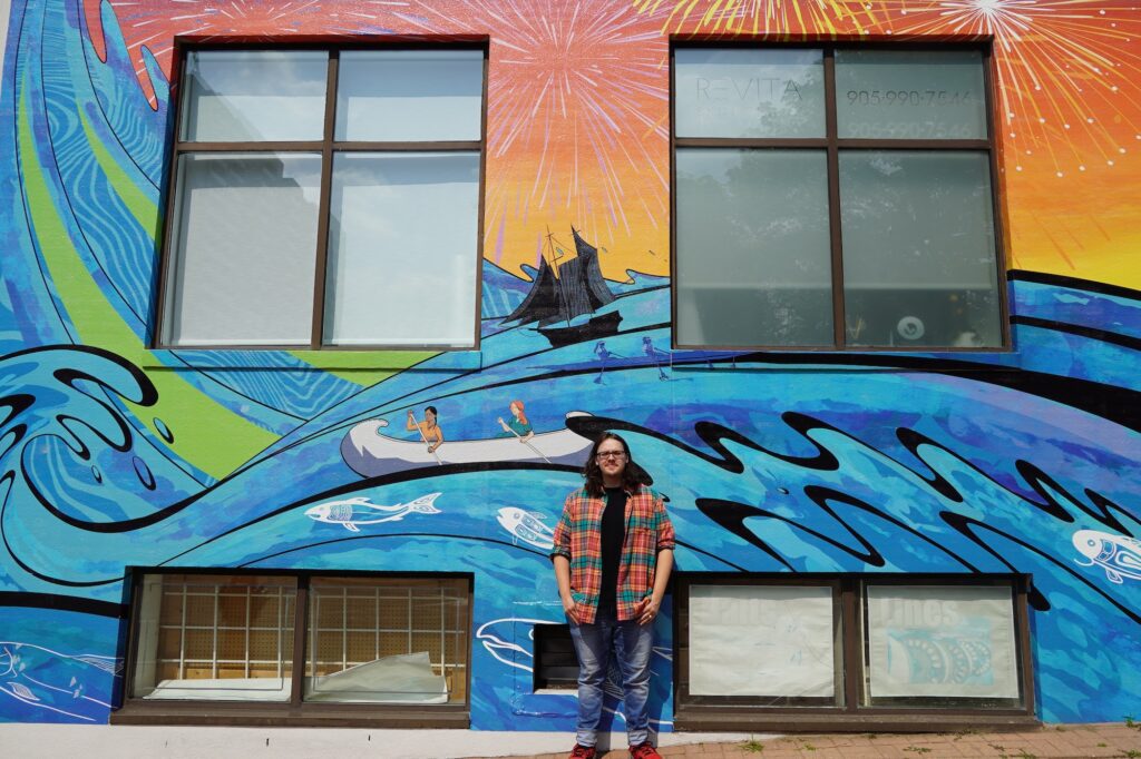 A man with glasses and a plaid shirt stands centred in front of a large mural. This part of the artwork depicts two people in a white canoe and a dark sailing ship navigating rolling blue and teal waves. Stylized white fish and a crane are also integrated into the water design. Above the waves, the sky is painted with bright orange and yellow sunbeams and white fireworks. The man is positioned between two large windows on the building's facade.