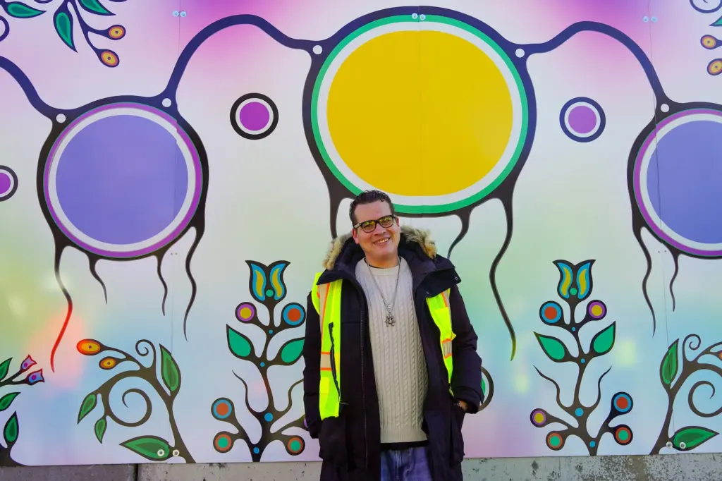 A man in a winter coat and safety vest smiling in front of a large, colourful Indigenous art mural featuring circular sun motifs and floral designs.
