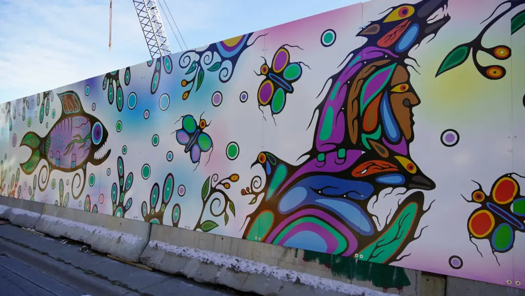 A digital illustration on construction hoarding on the side of a major street. The mural depicts fish, animals, and people in an Indigenous style.
