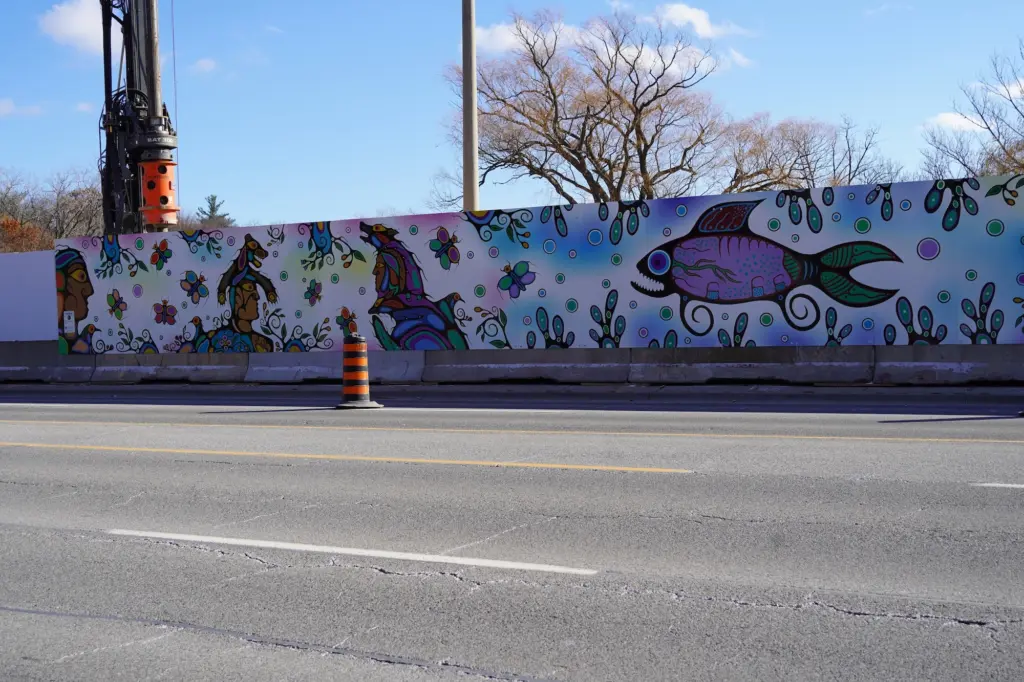 A digital illustration on construction hoarding on the side of a major street. The mural depicts fish, animals, and people in an Indigenous style.