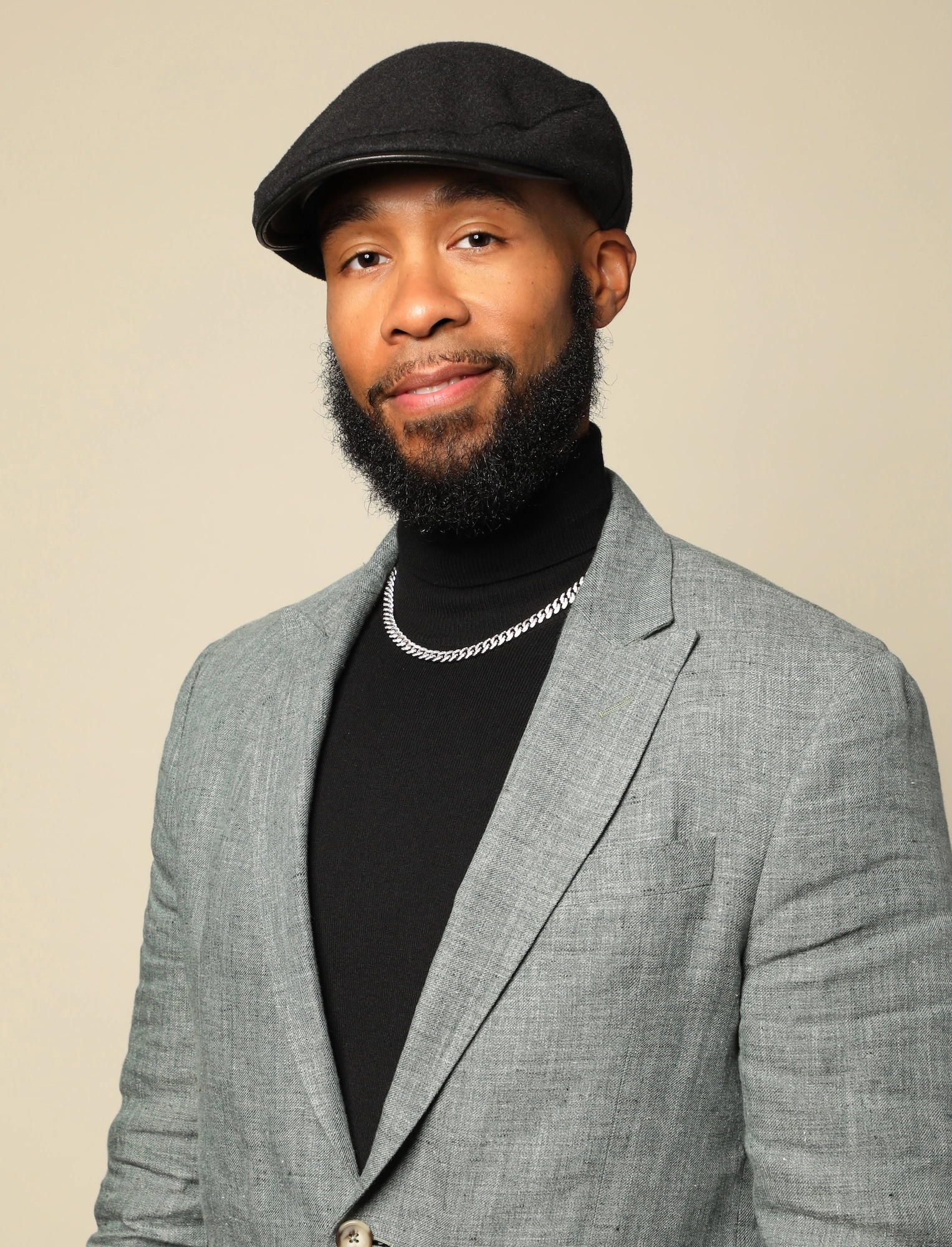Headshot of Matthew Testament Jones, who sits smiling against a beige background. He wears a black beret, black turtleneck with a silver chain, and a grey blazer.