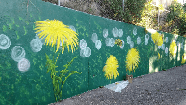 A hand-painted mural along a cement wall in a local park. The mural depicts dandelions and bumble bees against a dark green background.