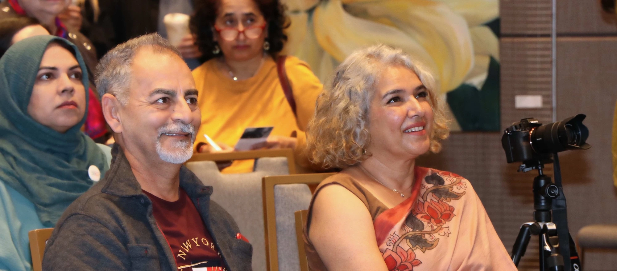 A man and a woman sitting in an audience and smiling. The woman is wearing a peach-coloured floral saree, and a camera on a tripod is visible in the foreground.