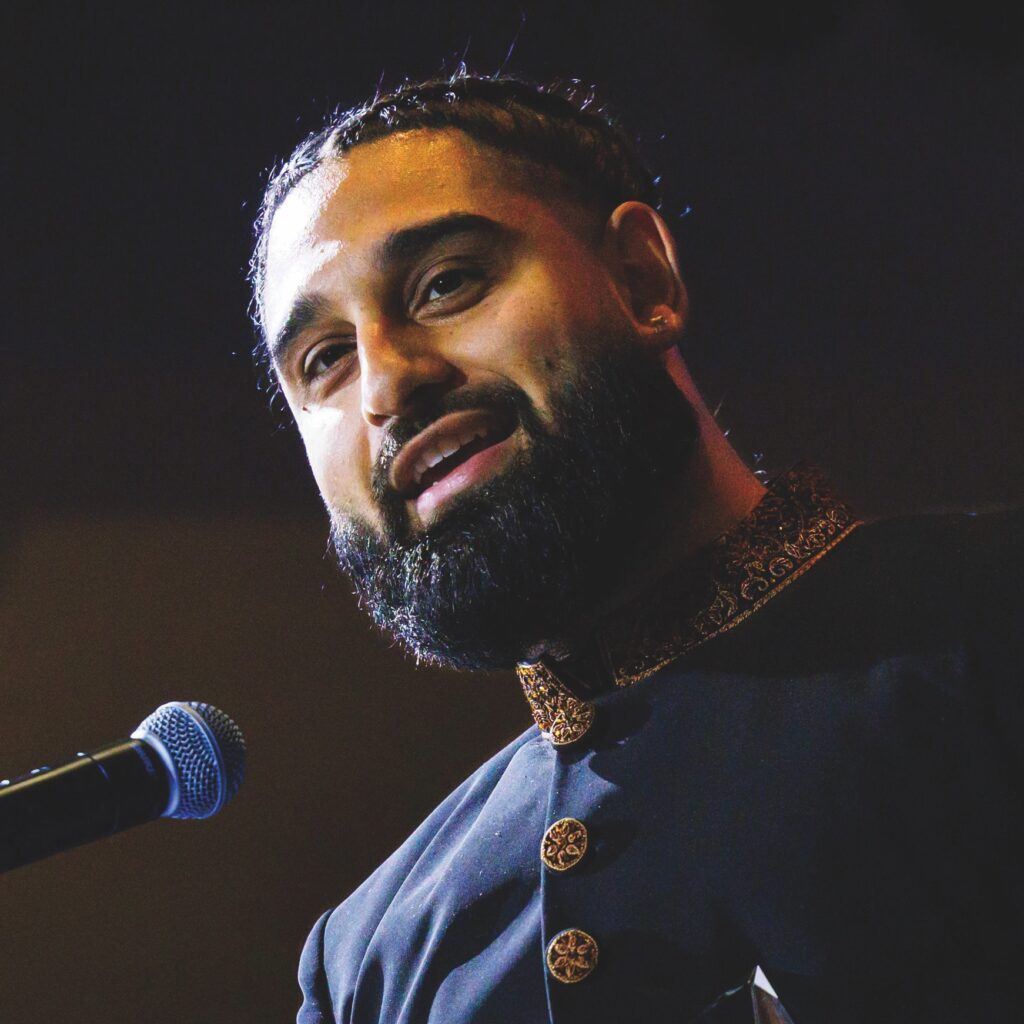 A medium close-up of Wali Shah, who wears a black collared suit with ornate gold trim and buttons. He performs a poetry piece in front of a microhpone on stage at the 2026 MARTY