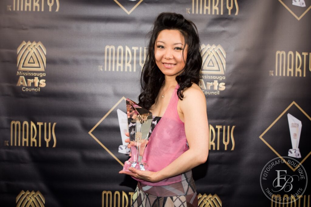 A woman in a pink dress stands in front of a photo backdrop with the words "MARTYS", the former MAC logo, and the award in gold on black. She holds a glass prism MARTY trophy.