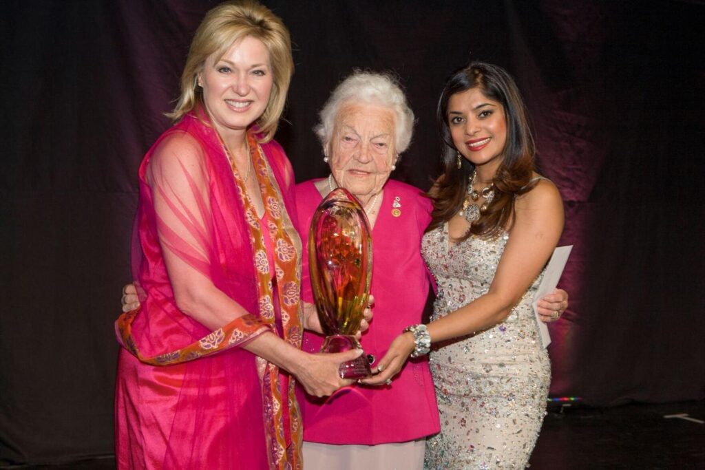 Three women stand together, with the two on the end holding the trophy for the elderly woman in the middle. The trophy is a large glass oval. 