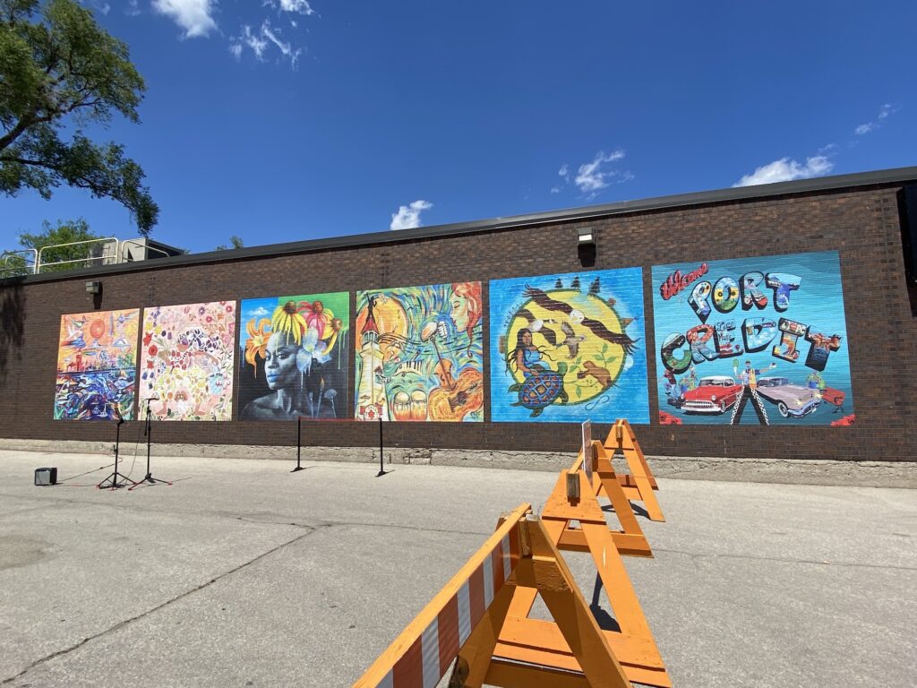 A brick wall featuring six vinyl murals on top, each with a distinct style and imagery. The building is situated in a parking lot. Bright orange barriers are visible, as well as a microphone and ribbon for cutting. 