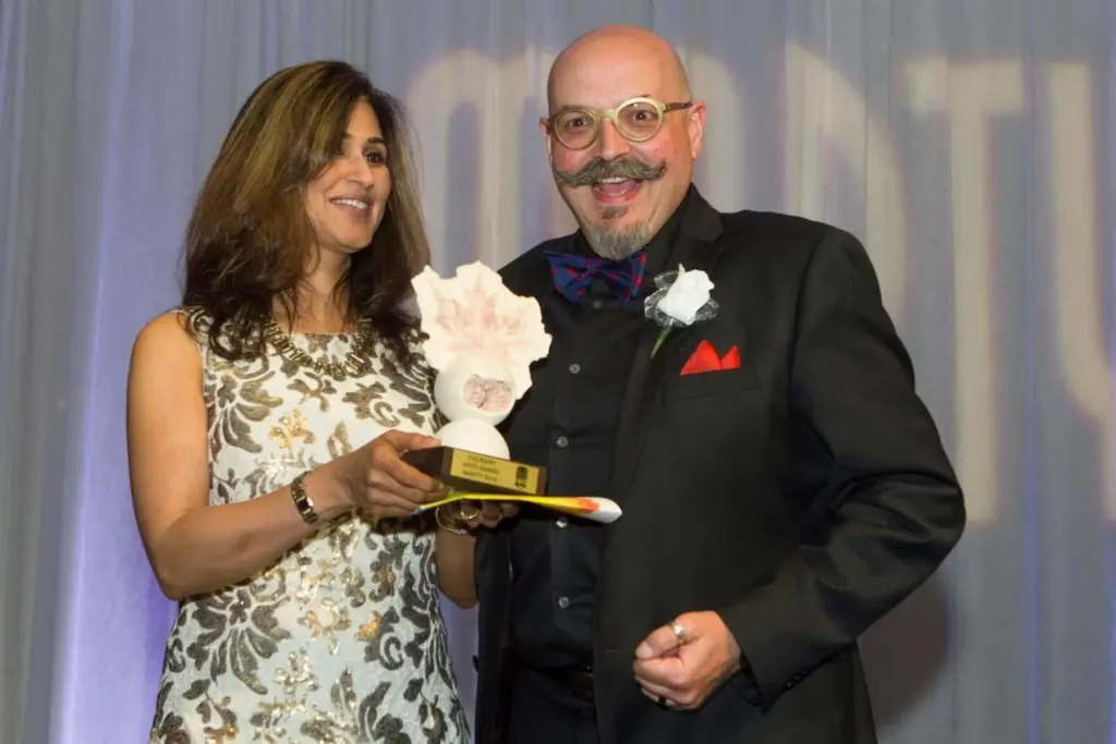 Two people pose on stage at the 2016 Mississauga Arts Awards. The woman on the left presents a stone award to the man smiling on the right.