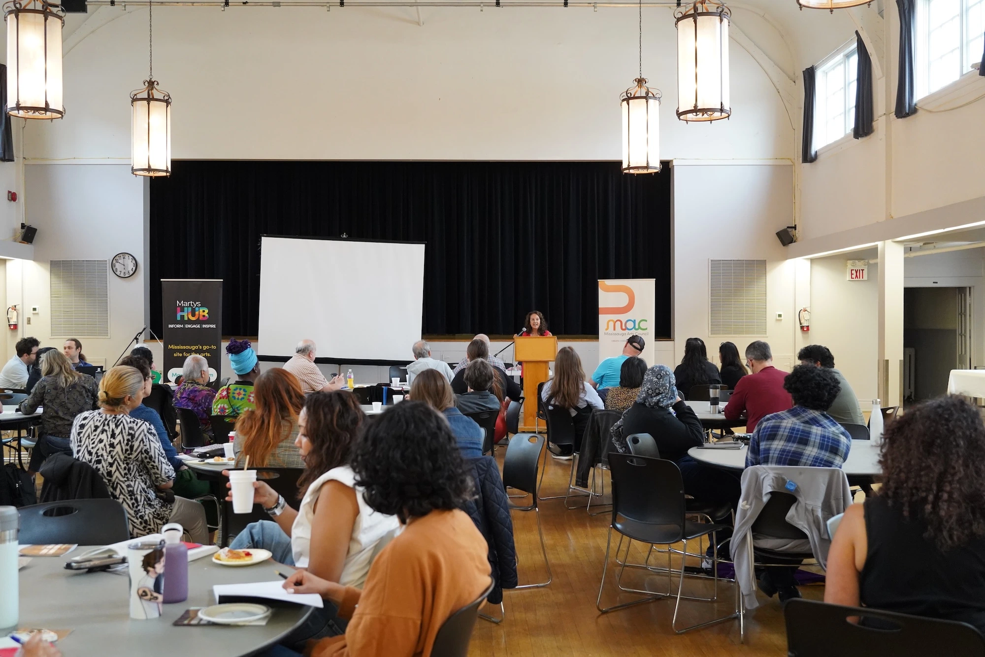 An overhead-style shot of a large, high-ceilinged hall with wooden floors and vintage hanging lanterns. A woman stands at a wooden lectern on a stage with a black curtain backdrop, addressing a large group of people seated at round tables. To her left is a large white projector screen.