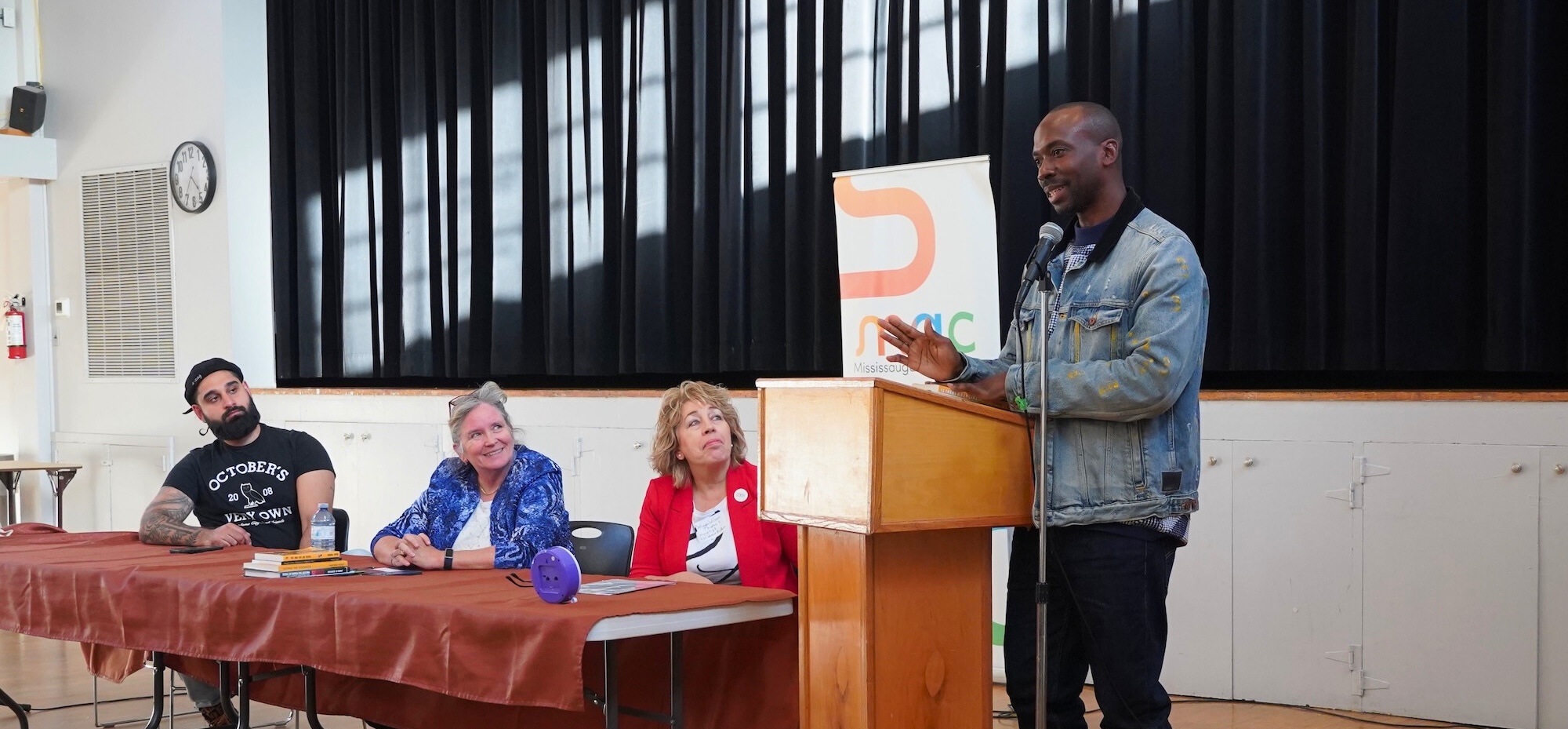 A man in a denim jacket stands at a wooden podium, speaking into a microphone during a panel event. Seated behind a long table covered in a burgundy cloth are three other panellists who are looking toward him and smiling. A "MAC" banner is visible in the background against a black stage curtain.
