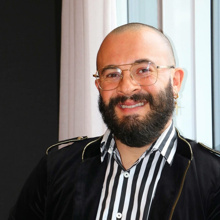 Headshot of Andrew Da Camara in a black jacket and white and black striped button down shirt against a window.