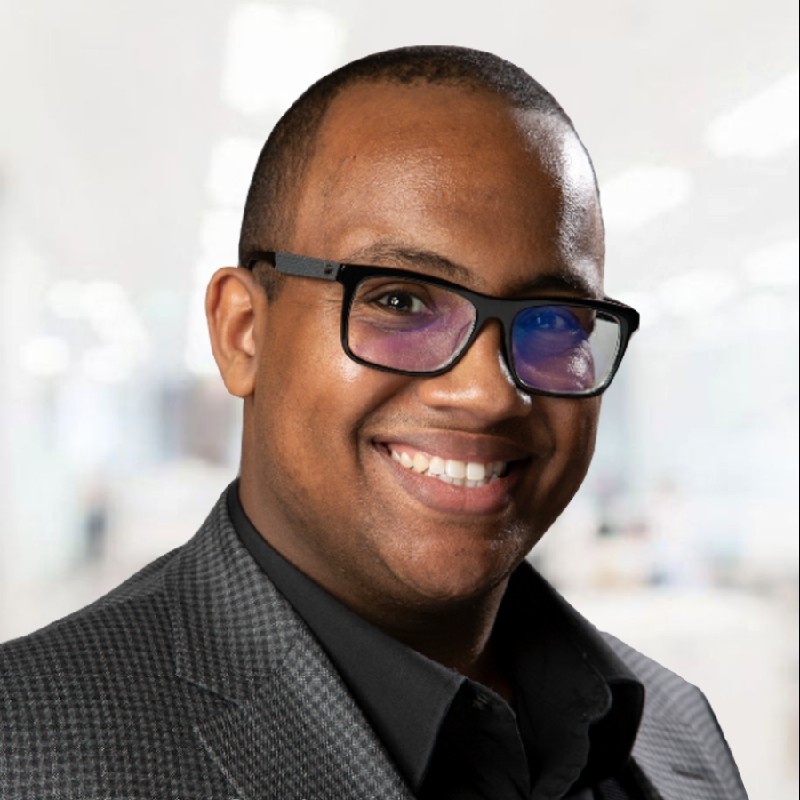 Headshot of Ayman Ashebir. He smiles in a black suit in front of a white backdrop.