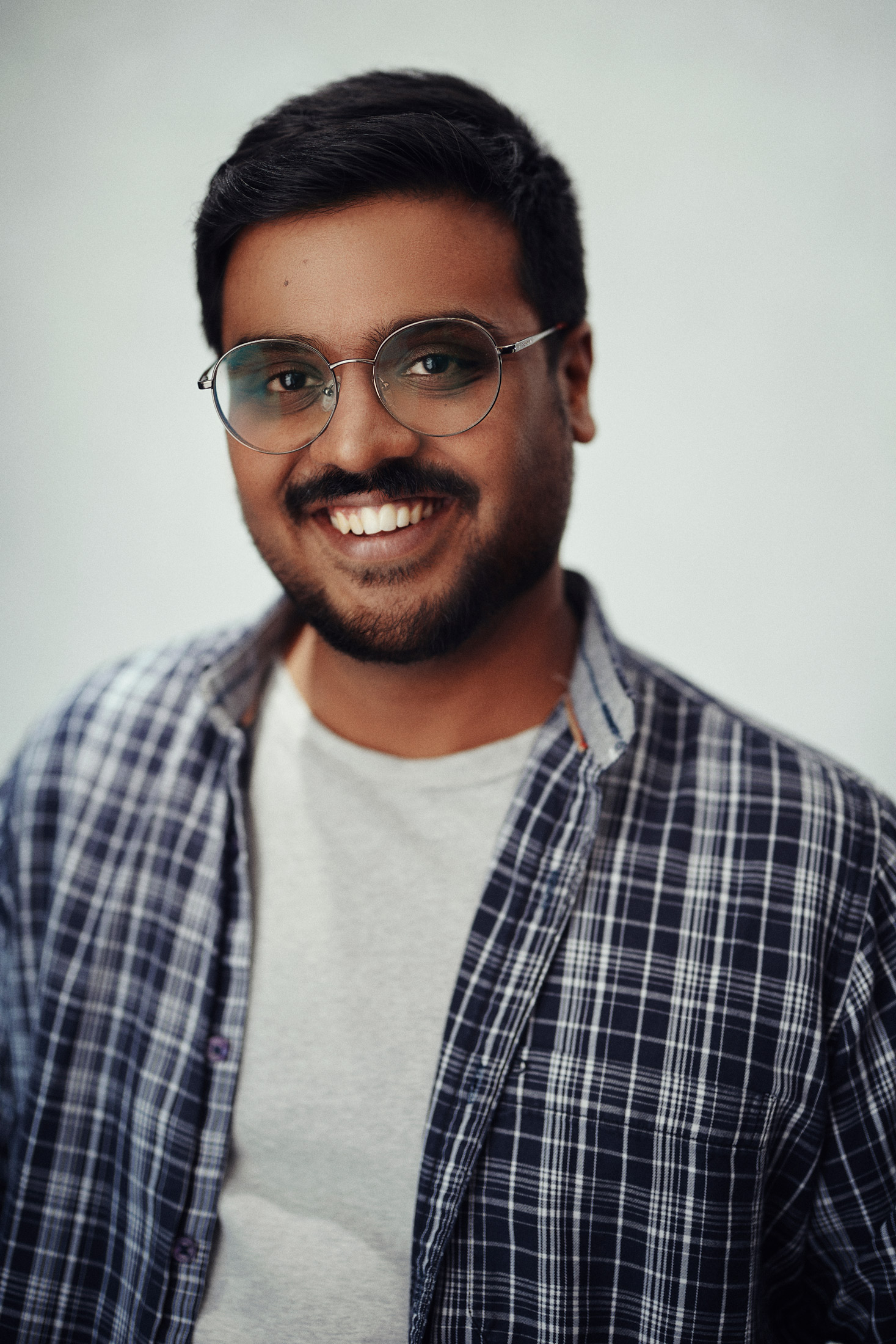 Headshot of Soma Narayanan, smiling wearing a plaid shirt and white t-shirt in front of a white backdrop.
