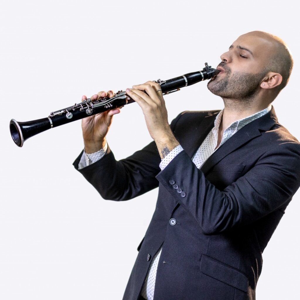 Photo of Majd Sukkar playing the clarinet. He wears a navy suit against a white backdrop.