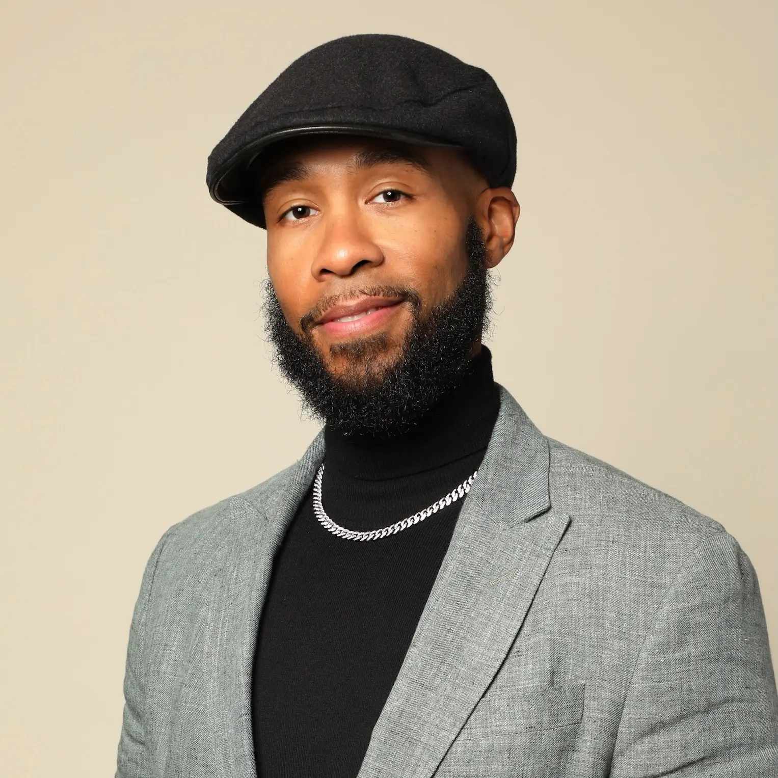 Headshot of Matthew Testament Jones, who sits smiling against a beige background. He wears a black beret, black turtleneck with a silver chain, and a grey blazer.