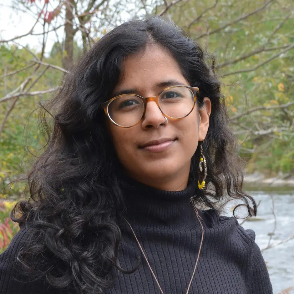 Headshot of Manahil Bandukwala. She wears a black turtleneck, turtleshell glasses, and a long necklace as well as beaded earrings. She stands in front of a forest backdrop with a river visible in the bottom right.