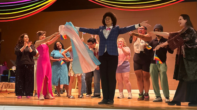 A group of joyful performers celebrate together on a stage beneath rainbow streamers. In the centre, a person in a blue blazer and black trousers stands with arms wide open. Other individuals surround them, smiling, clapping, and holding Pride and transgender flags.