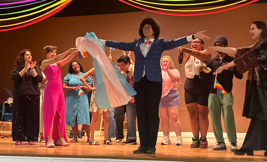 A group of joyful performers celebrate together on a stage beneath rainbow streamers. In the centre, a person in a blue blazer and black trousers stands with arms wide open. Other individuals surround them, smiling, clapping, and holding Pride and transgender flags.