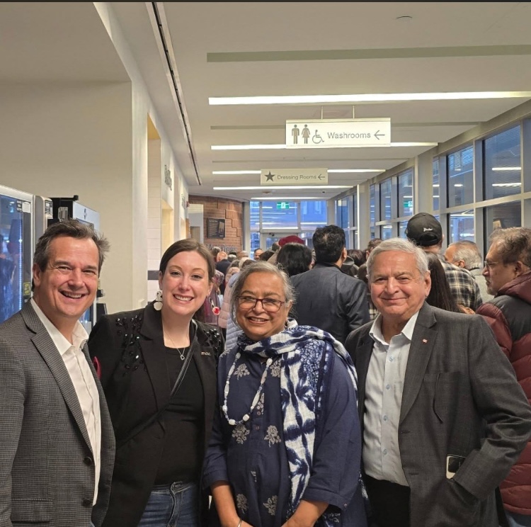 Four people smiling together for a photo in a brightly lit indoor hallway in front of a large crowd.