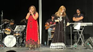 A live musical performance on a stage with a black backdrop. Two women in formal dresses sing into microphones at the front, supported by a live band including a drummer, a bassist, and a keyboardist.