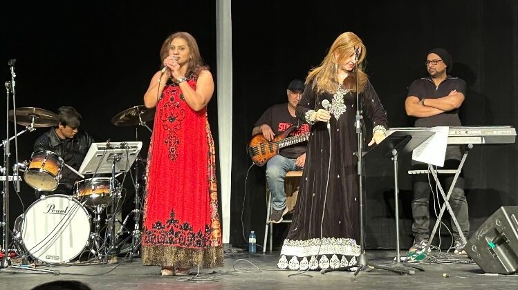 A live musical performance on a stage with a black backdrop. Two women in formal dresses sing into microphones at the front, supported by a live band including a drummer, a bassist, and a keyboardist.