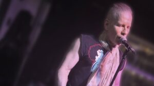 A musician performs live on a stage illuminated by moody purple light. He wears a black sleeveless graphic tee, dark pants, and a light-colored scarf wrapped around his neck while singing into a microphone and playing a black electric guitar.