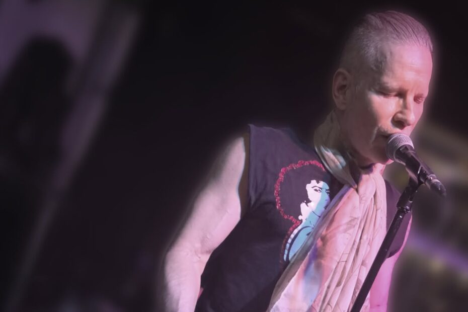 A musician performs live on a stage illuminated by moody purple light. He wears a black sleeveless graphic tee, dark pants, and a light-colored scarf wrapped around his neck while singing into a microphone and playing a black electric guitar.
