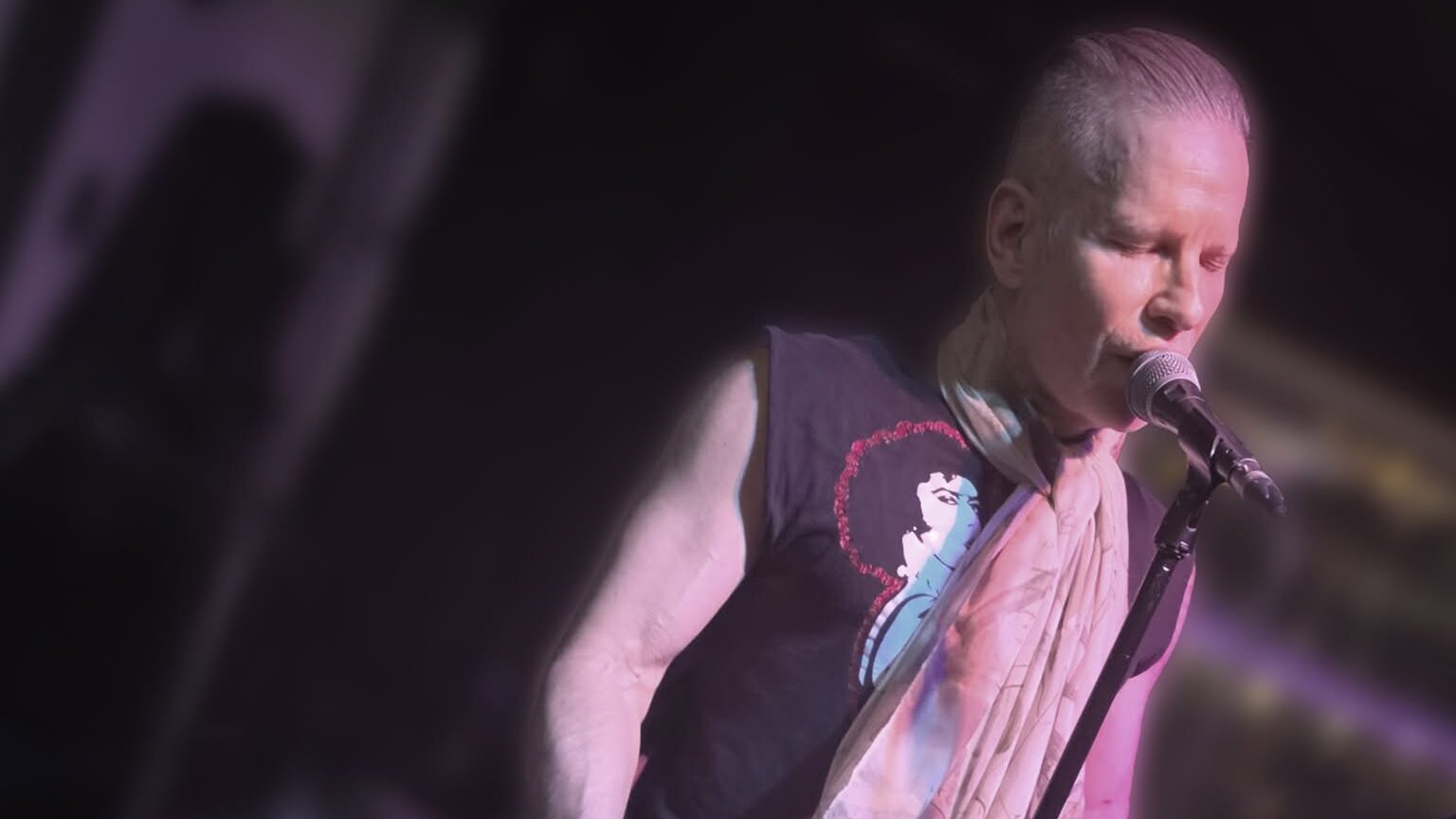 A musician performs live on a stage illuminated by moody purple light. He wears a black sleeveless graphic tee, dark pants, and a light-colored scarf wrapped around his neck while singing into a microphone and playing a black electric guitar.