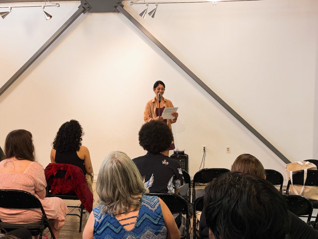 A woman standing at a microphone reading from papers to a seated audience during an indoor event.