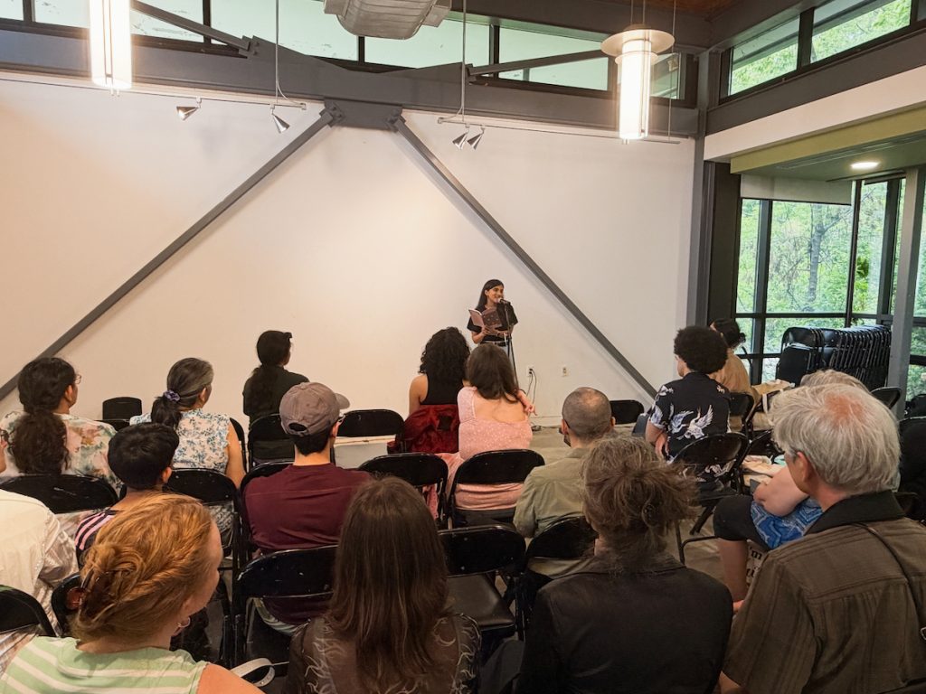 Wide shot from behind an audience looking toward a speaker presenting at a microphone in a brightly lit room with large windows.