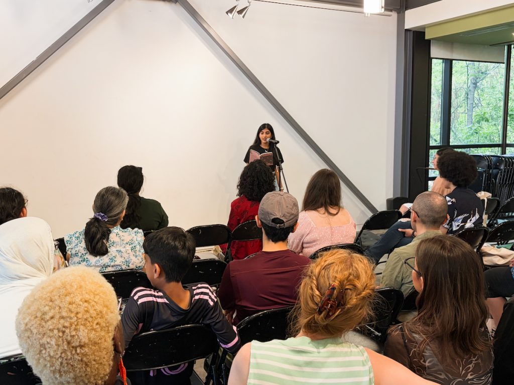 Audience members seated in black folding chairs facing a speaker reading from a book at the front of the room.