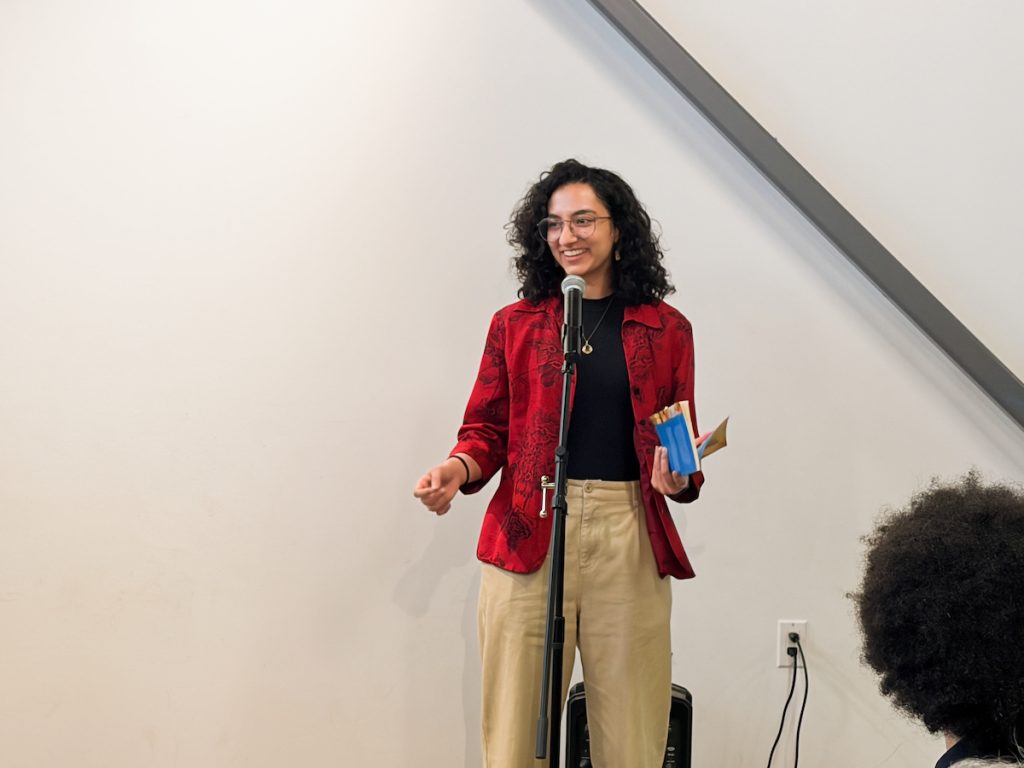 A person with curly hair, wearing glasses and a red patterned jacket, speaking into a microphone while holding a small book.
