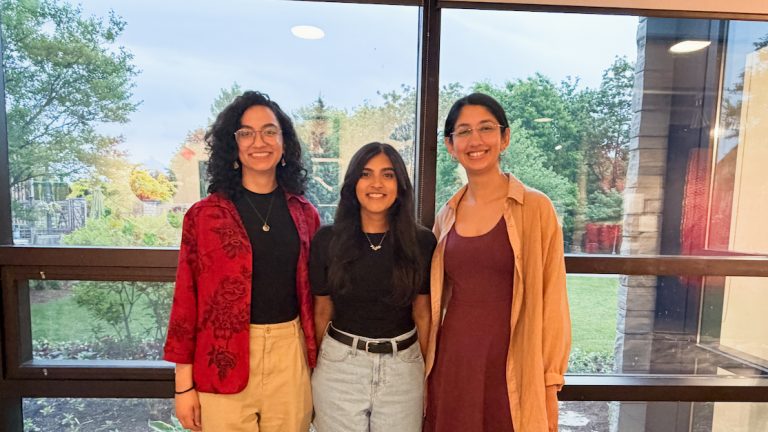Three smiling people standing side-by-side posing for a photo in front of a large window.
