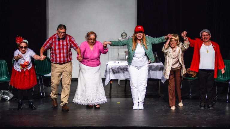 Six performers holding hands and bowing in a row on a stage at the conclusion of the play "Fue Sin Querer Queriendo." They are dressed in colorful, expressive costumes, including a red baseball cap, a pink cardigan, a red blazer, and a checkered shirt.