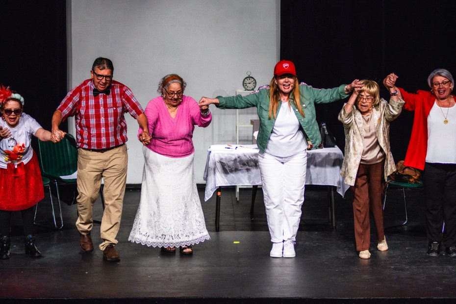 Six performers holding hands and bowing in a row on a stage at the conclusion of the play "Fue Sin Querer Queriendo." They are dressed in colorful, expressive costumes, including a red baseball cap, a pink cardigan, a red blazer, and a checkered shirt.