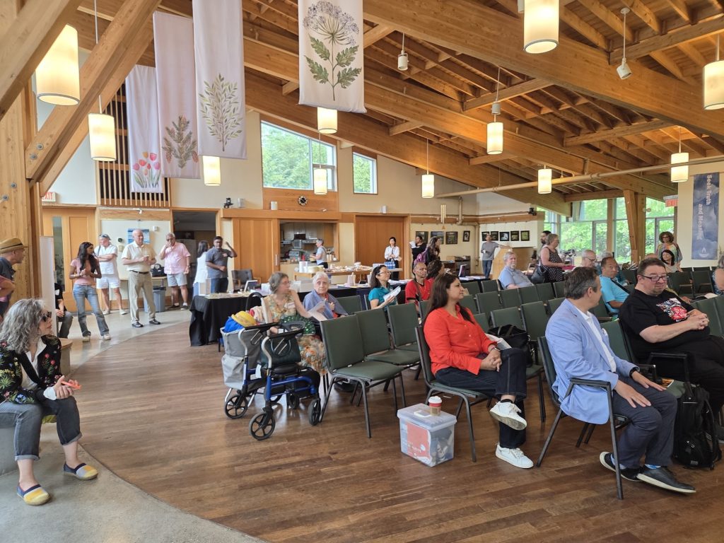 People seated in rows of chairs and standing inside a large wooden hall with high ceilings, attending an event.
