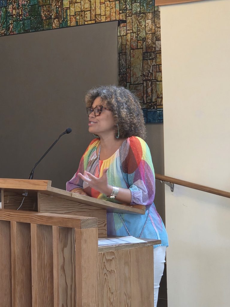 A woman with curly hair wearing a colourful blouse speaking into a microphone at a wooden podium.