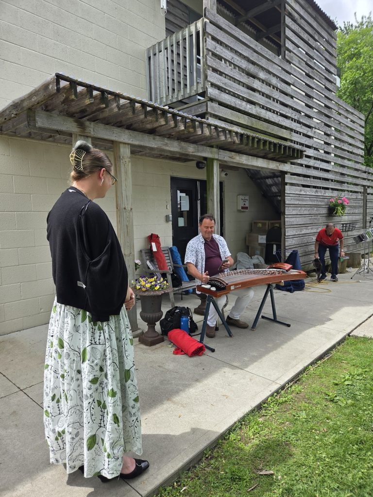 A man playing a traditional stringed zither instrument outdoors on a patio while a woman looks on.