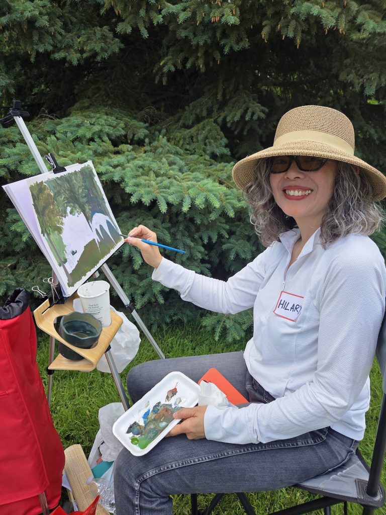 A woman wearing a straw hat and glasses smiling while painting on a canvas outdoors in front of evergreen trees.