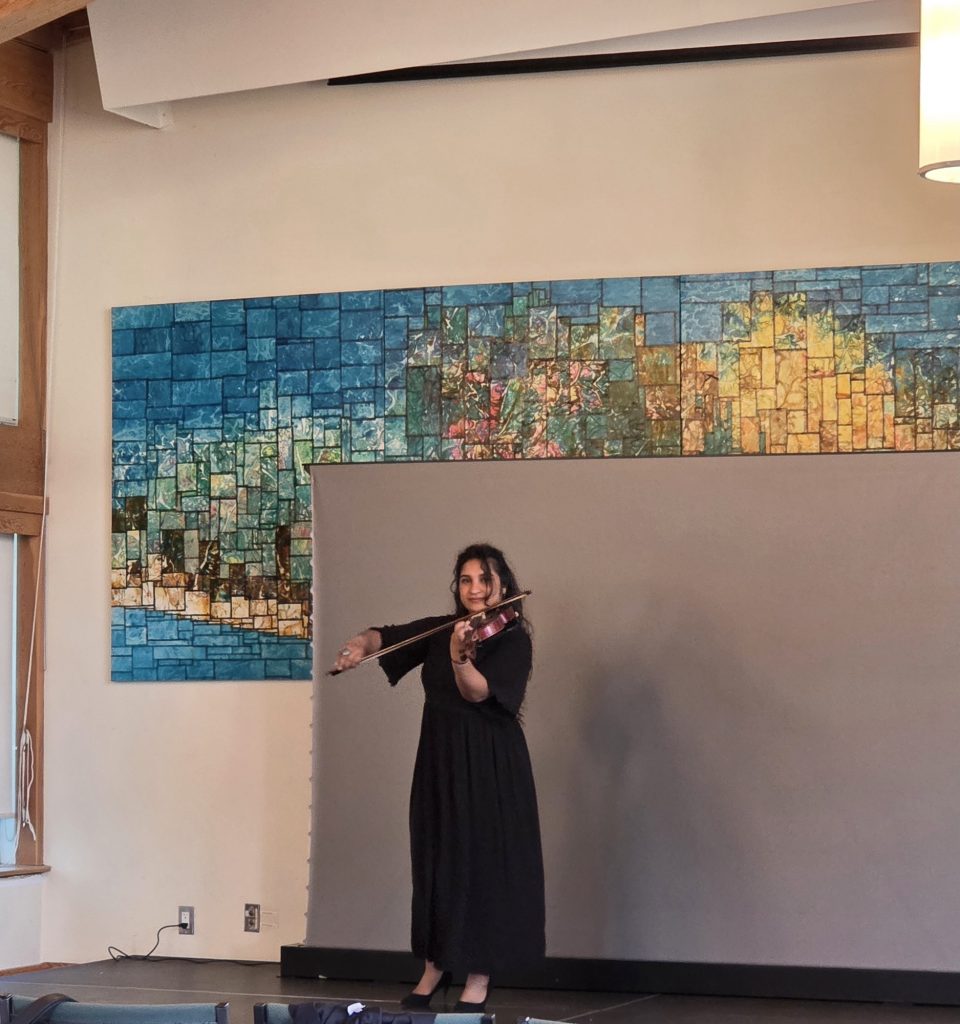 A woman in a black dress performing on the violin in front of a large, colourful mosaic wall feature.