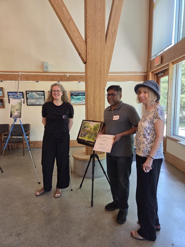 A man holding an award certificate posing with two women next to a watercolour painting on an easel.
