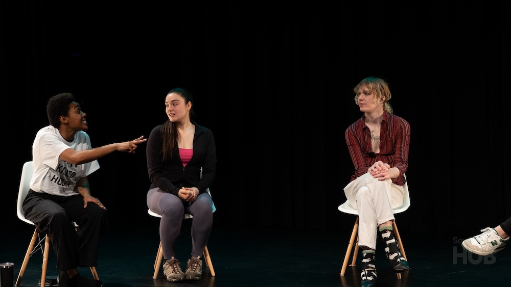 A close-up of three people sitting on stage in white chairs. The person on the left gestures and speaks animatedly toward the woman in the centre, who listens attentively, while the person on the right looks on calmly with hands clasped.