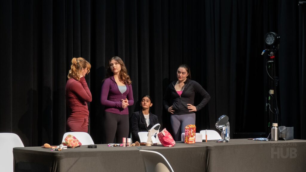 Four young women are gathered behind a long table covered with a black tablecloth. The table is scattered with makeup bags, cosmetics, and a small mirror.