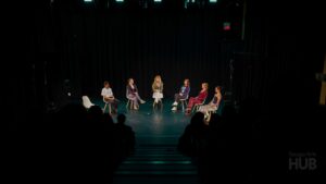 A wide shot of a theatre stage with six people seated in white chairs arranged in a semi-circle, participating in a panel or discussion. The stage is dimly lit with a black curtain backdrop, and the dark silhouettes of an audience are visible in the foreground.