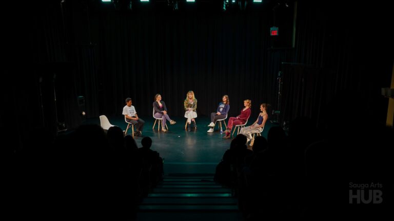 A wide shot of a theatre stage with six people seated in white chairs arranged in a semi-circle, participating in a panel or discussion. The stage is dimly lit with a black curtain backdrop, and the dark silhouettes of an audience are visible in the foreground.