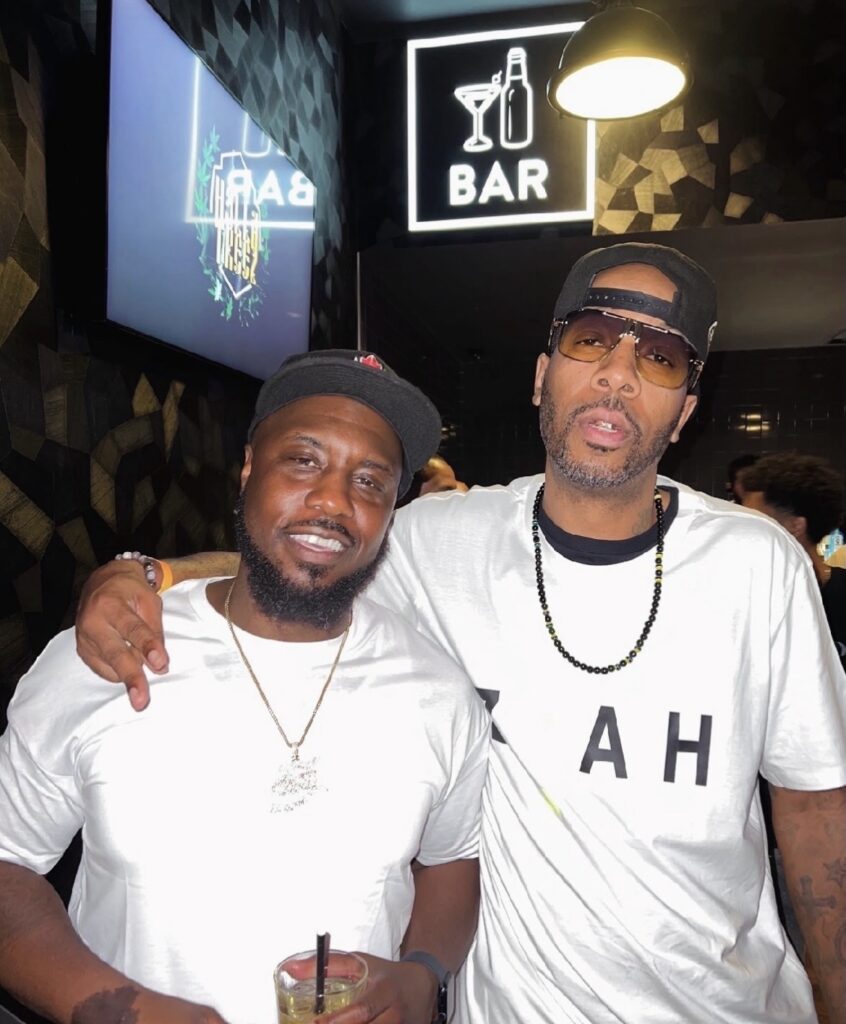 A close-up photo of two men smiling. The man on the left wears a white t-shirt and a black cap while holding a drink; the man on the right wears a white graphic tee, sunglasses, and a backwards cap with his arm around the other's shoulder.