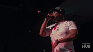 A close-up, low-angle shot of a man in a white t-shirt and a black baseball cap rapping passionately into a microphone under stage lighting.