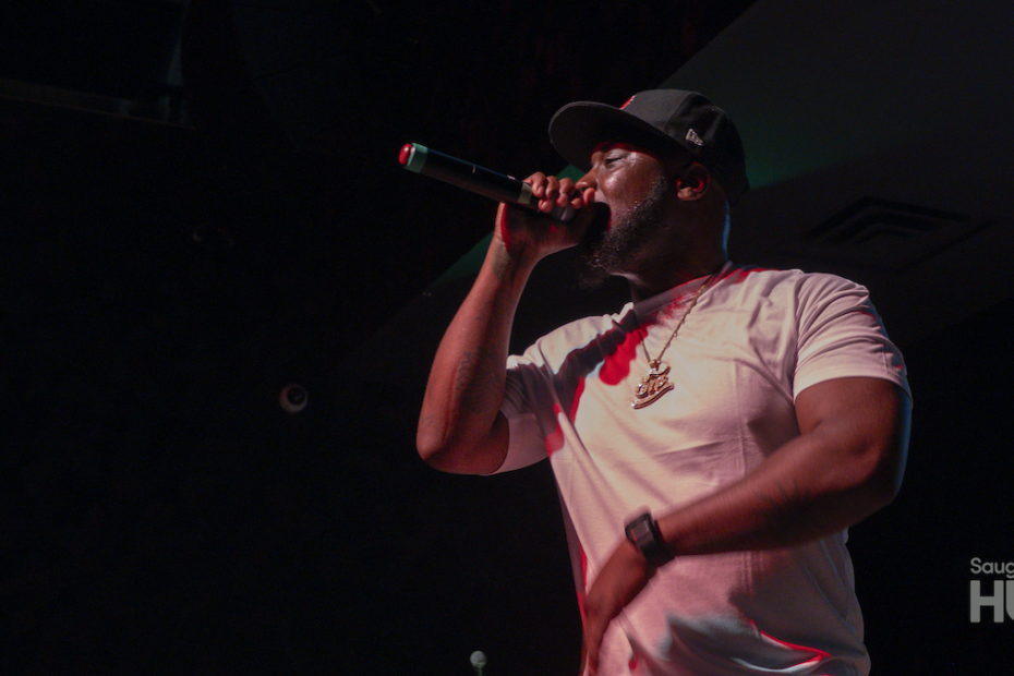 A close-up, low-angle shot of a man in a white t-shirt and a black baseball cap rapping passionately into a microphone under stage lighting.