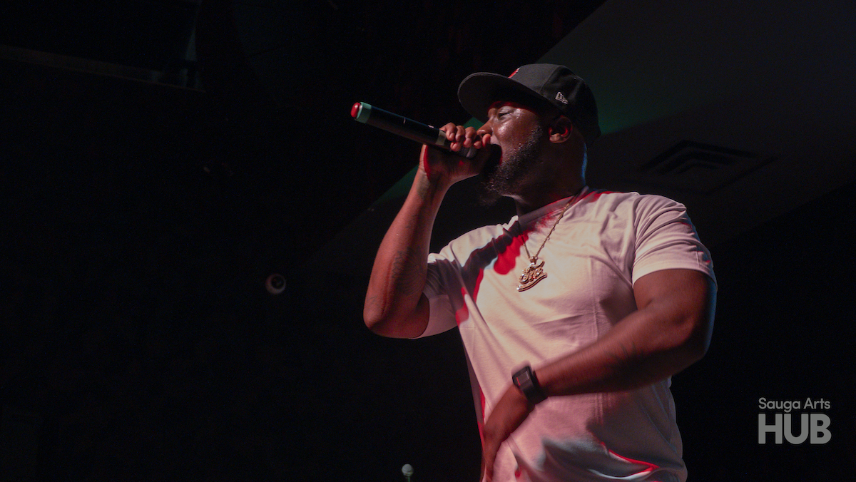 A close-up, low-angle shot of a man in a white t-shirt and a black baseball cap rapping passionately into a microphone under stage lighting.