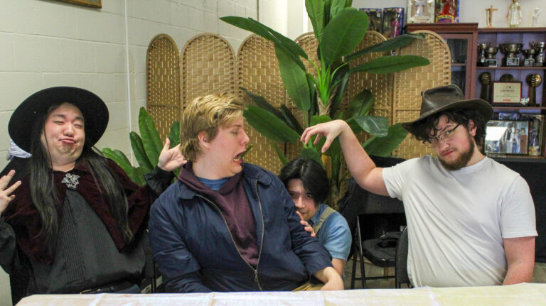 A group of four people sitting behind a table, playfully posing with exaggerated expressions. The person on the left is making a funny face, while the person on the right in a cowboy hat mimics a monster claw with his hand over another person's head.